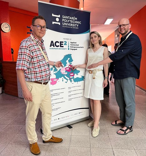 Trzy osoby stoją przy roll-upie Santarém Polytechnic University z logo ACE² European University i mapą Europy; uśmiechnięte, wskazują palcem wybrane miejsce na mapie w jasnym korytarzu z czerwonymi ścianami.