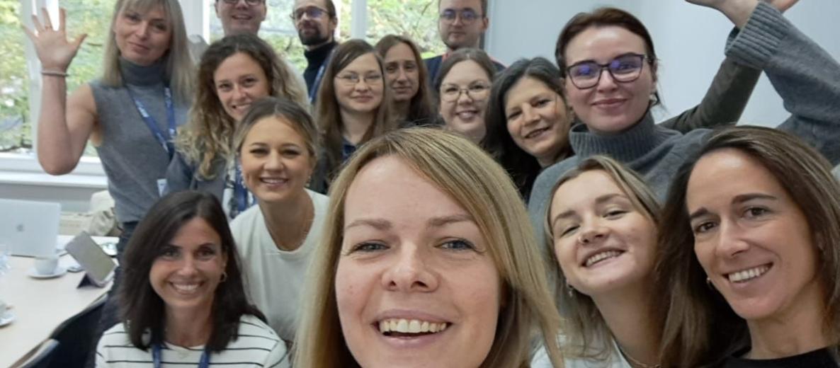 Grupowe selfie w jasnej sali z dużymi oknami; kilkanaście uśmiechniętych osób, część macha do aparatu podczas warsztatów.