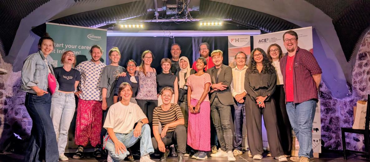 Duża, uśmiechnięta grupa ok. 20 osób pozuje na scenie w kamiennej sali z łukowym sufitem; w tle roll-upy partnerów, m.in. Infineon i ACE² European University, sceniczne światła i sprzęt.