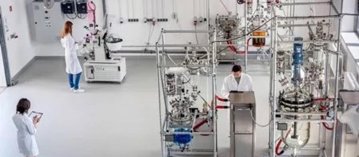 Laboratorium z trzema osobami w fartuchach; aparatura chemiczna z reaktorami i szkłem laboratoryjnym w jasnym, sterylnym wnętrzu.