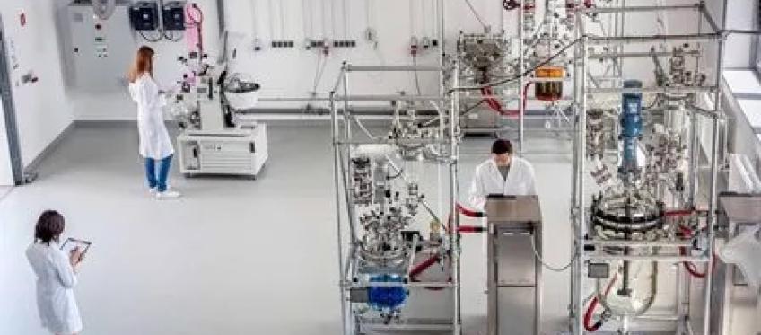 Laboratorium z trzema osobami w fartuchach; aparatura chemiczna z reaktorami i szkłem laboratoryjnym w jasnym, sterylnym wnętrzu.