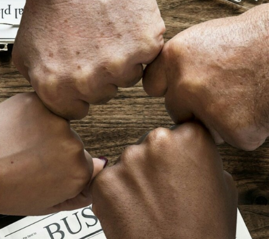 Four colleagues bump fists over a wooden desk with a laptop, documents, and coffee—symbolizing teamwork and support.
