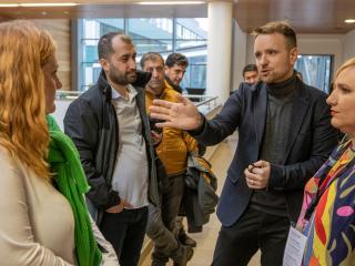 Rozmowa w korytarzu: mężczyzna w marynarce gestykuluje, wokół stoi kilka osób słuchających.