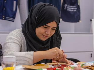 Młoda kobieta w hijabie dekoruje pierniki przy stole, w tle materiały plastyczne.