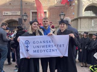 Trzy osoby przed zabytkowym budynkiem trzymają baner „GDAŃSKI UNIWERSYTET MEDYCZNY”; dwie w togach akademickich.