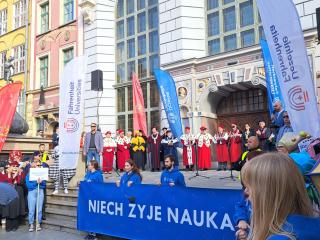 Uroczystość na Długim Targu; naukowcy w togach na scenie, a przed nimi studenci w niebieskich bluzach z banerem „NIECH ŻYJE NAUKA”.