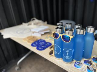 Stół z gadżetami promocyjnymi: niebieskie butelki wielorazowe z logo USS GUMed, okulary i naklejki.