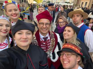 Grupowe selfie w strojach akademickich i ludowych podczas miejskiej uroczystości; wszyscy uśmiechnięci.